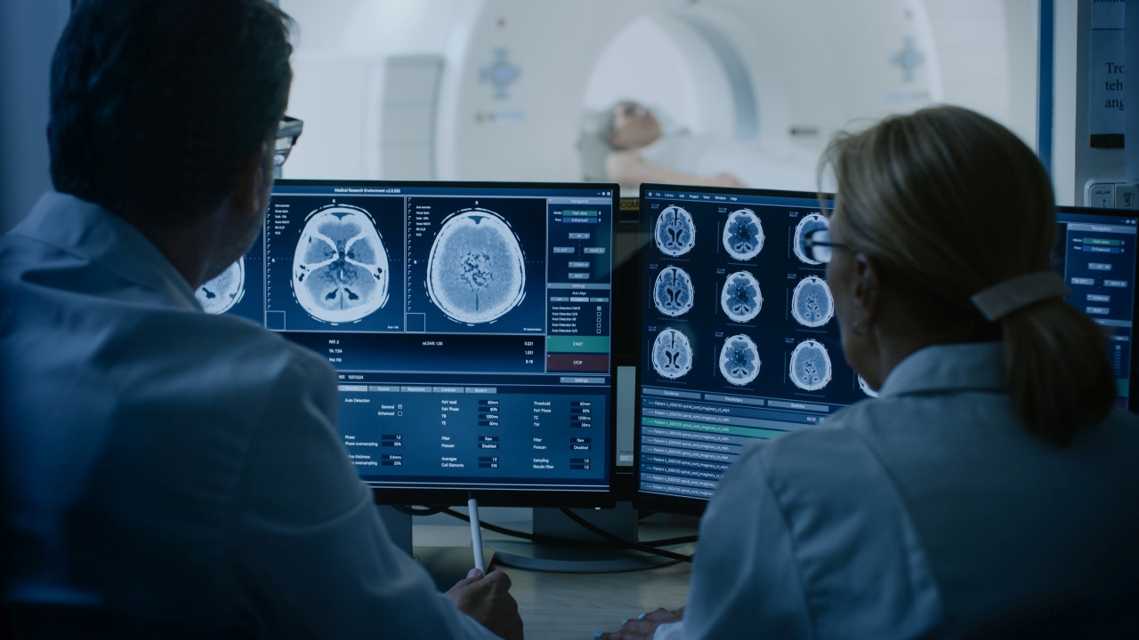 In Control Room Doctor and Radiologist Discuss Diagnosis while Watching Procedure and Monitors Showing Brain Scans Results, In the Background Patient Undergoes MRI or CT Scan Procedure