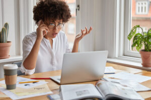 Puzzled shocked indignant female employee shruggs shoulders in frustration, looks at laptop computer