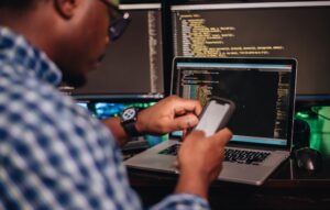 African American man sitting at computer developer software while using smartphone